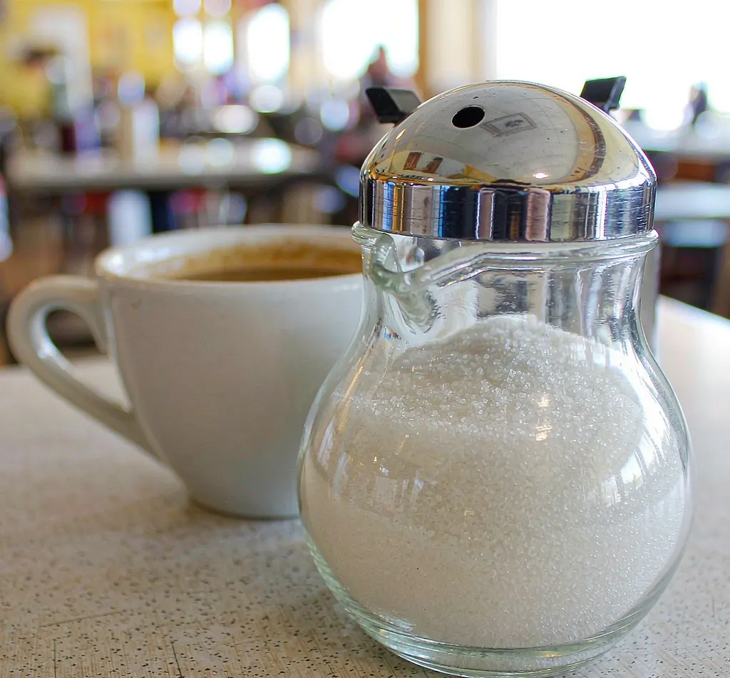 Glass sugar pourer bottle with chrome dome lid beside a coffee cup